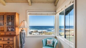 A bedroom with a large window overlooking the ocean, a wooden cabinet, and a teal chair with a starfish pillow. - 3976 Beach House on Asilomar~ Oceanfront View Home (Pacific Grove)