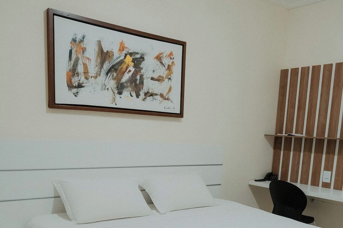 Een hotelkamer met een groot bed, een nachtkastje, een bureau, een stoel, een raam met jaloezieën en een schilderij aan de muur.