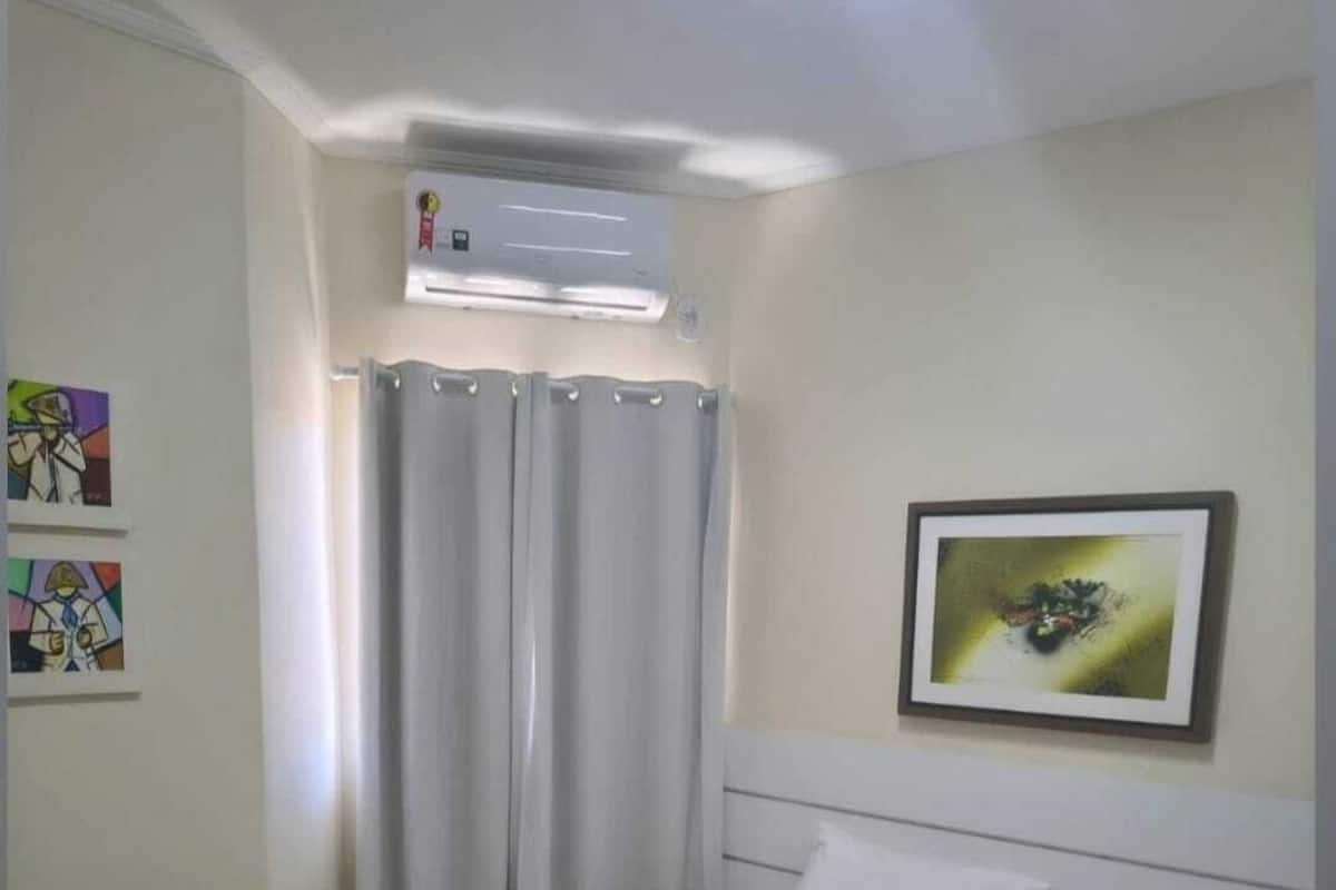 Een hotelkamer met een bed, nachtkastje, airconditioning, gordijn en een lijst met een foto aan de muur.