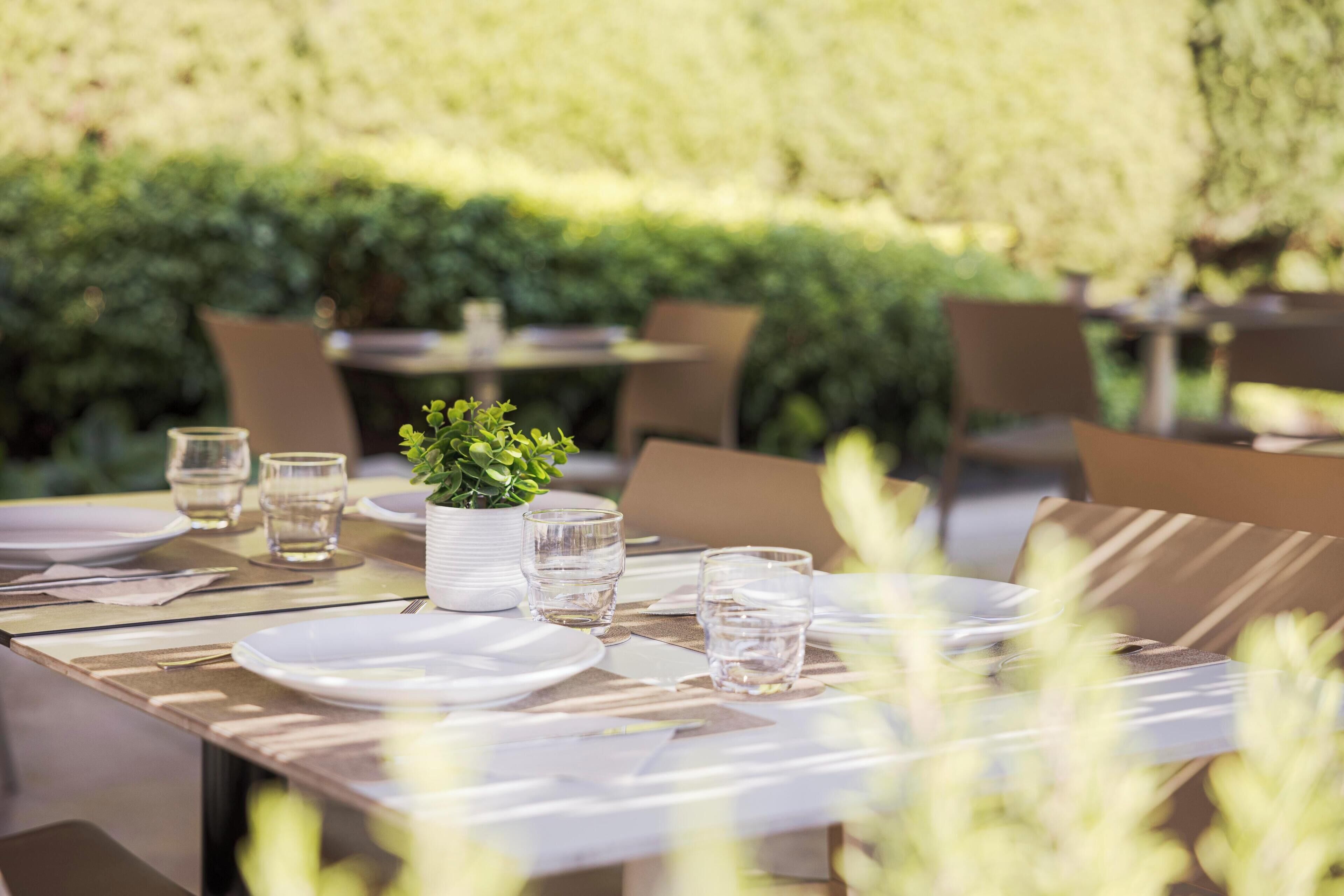 An outdoor dining area with tables set for two, featuring clear glassware and a potted plant centerpiece.