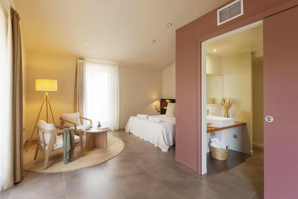 Ein modernes Hotelzimmer mit einem Bett, einer Sitzecke mit Tisch und Stühlen, einem Badezimmer mit Waschbecken und Spiegel sowie einem großen Fenster mit Vorhängen.