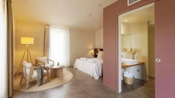 Une chambre d’hôtel moderne comprenant un lit, un coin salon avec une table et des chaises, une salle de bain avec un lavabo et un miroir, et une grande fenêtre avec des rideaux.