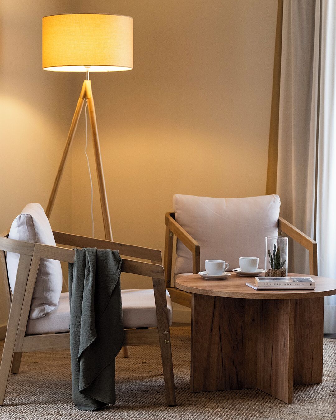 A modern hotel room with a wooden floor, a round wooden coffee table, a white armchair, a wooden chair with a cushion, a floor lamp, and a small plant on the table.
