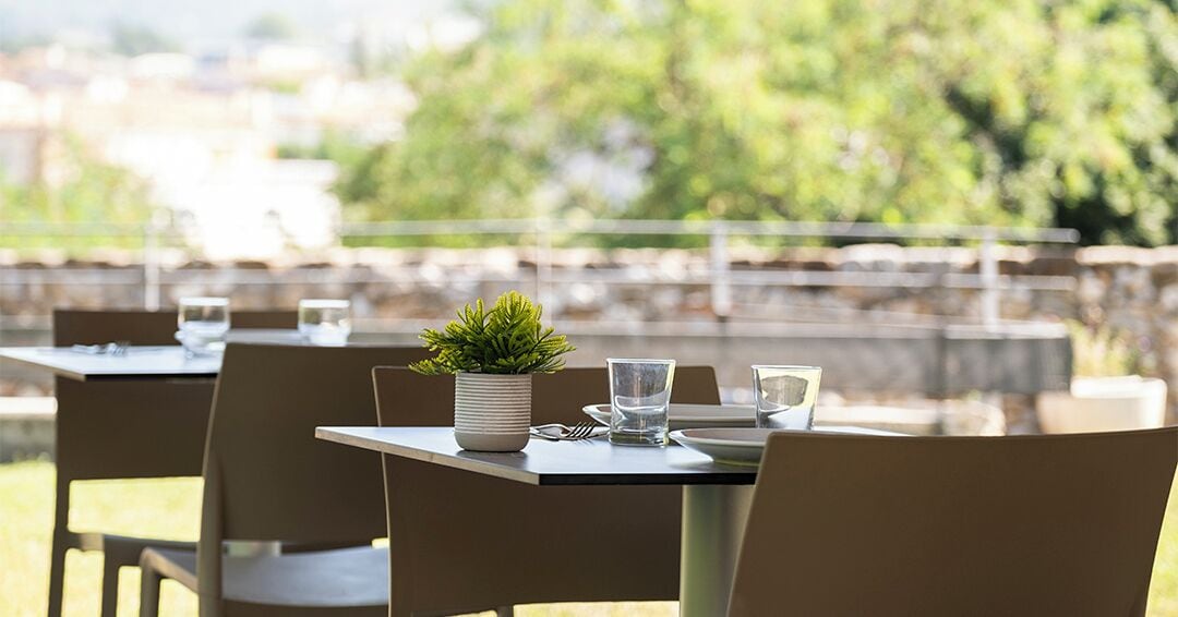 Un espacio de comedor al aire libre con mesas dispuestas para dos, cada una con una planta en maceta, vasos y cubiertos.