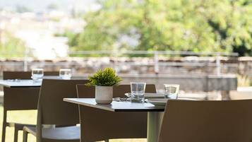 Un espacio de comedor al aire libre con mesas dispuestas para dos, cada una con una planta en maceta, vasos y cubiertos.