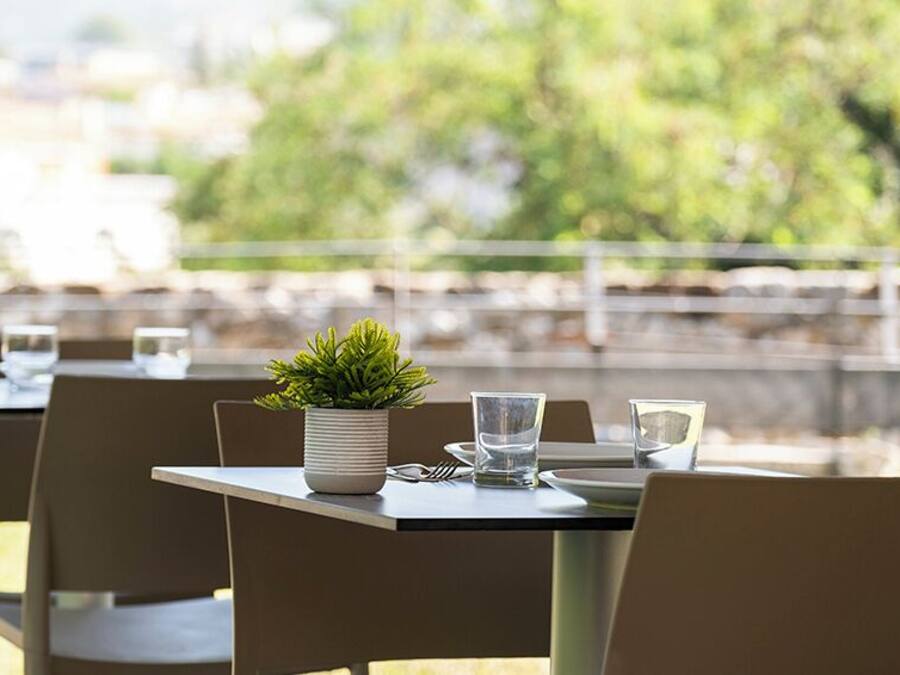 Un espacio de comedor al aire libre con mesas dispuestas para dos, cada una con una planta en maceta, vasos y cubiertos.