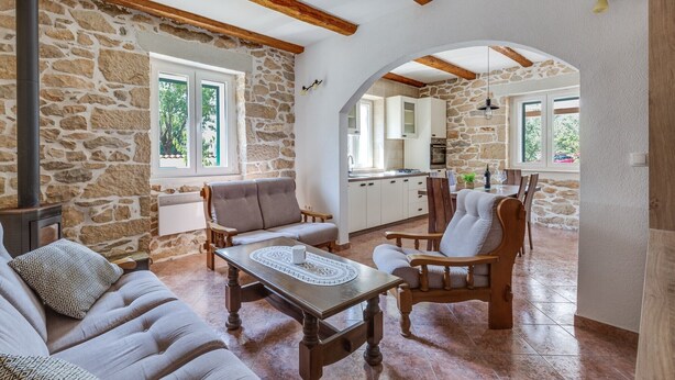 A cozy living room with stone walls, wooden beams, a fireplace, and a view into the kitchen. - Haus Zdravko in Prkos  - Haus für 4 Personen (Prkos)