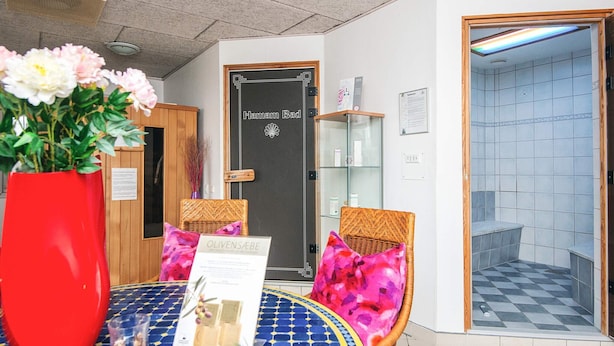 A reception area with a check-in counter, a large red vase with flowers, wicker chairs with colorful cushions, and a tiled floor.