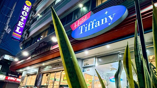 Um hotel com um letreiro Tiffany bem visível, uma entrada evidente e vista da rua com um carro estacionado.