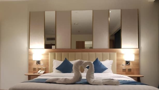 A hotel room with a large bed, a headboard with a mirror, two bedside lamps, and a swan-shaped towel decoration. - ALHYAT Hotel (Jeddah)