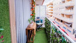 A narrow balcony with artificial grass, a wooden table, and chairs, surrounded by greenery and a view of a building with balconies.