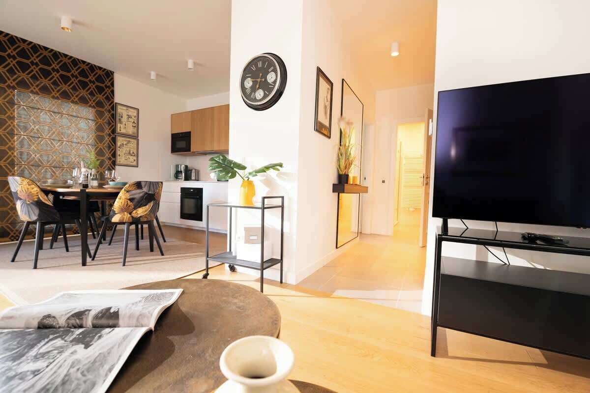 A modern living room with a flat-screen TV, a dining area with a table and chairs, and a kitchen in the background.