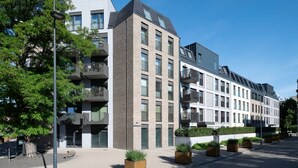 Un edificio residencial moderno de varios pisos, con balcones, techo plano y un patio empedrado con jardineras.