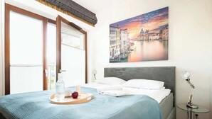 Una habitación de hotel con una cama, un cuadro de un paisaje urbano, una mesita de noche con una lámpara y una ventana con vistas al exterior.