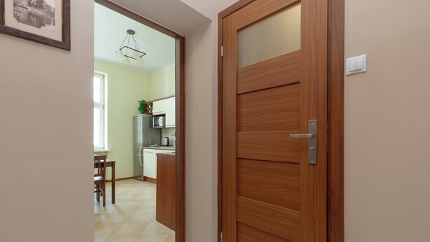 A wooden door with a modern handle, leading to a kitchen with white cabinets and a dining area with a table and chairs.