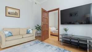 A living room with a beige sofa, a coffee table, a rug, a wooden door, a bed, and a TV mounted on the wall.