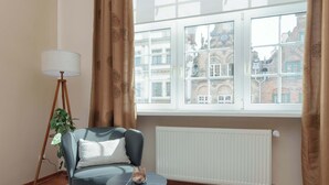 A room with a blue armchair, a small round table, a floor lamp, and a view of a building through a window with curtains.