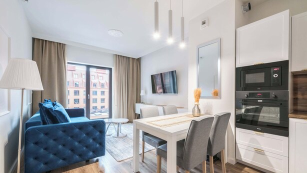 A modern hotel room with a blue sofa, dining table, chairs, a TV, and a kitchenette with a microwave and oven.