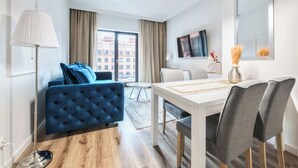 A modern hotel room with a blue sofa, a dining table with chairs, a large window with curtains, and a flat-screen TV mounted on the wall.