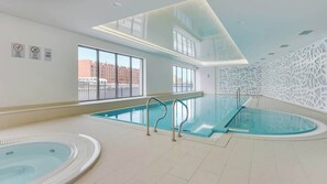 A modern indoor swimming pool with clear glass walls, a view of buildings outside, and a decorative white wall feature.