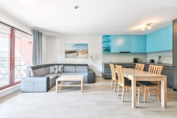 A modern living room with a sofa and a dining table, a kitchen with blue cabinets, and a large window.