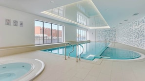 A modern indoor swimming pool with clear glass walls, a view of buildings outside, and a decorative wall on one side.