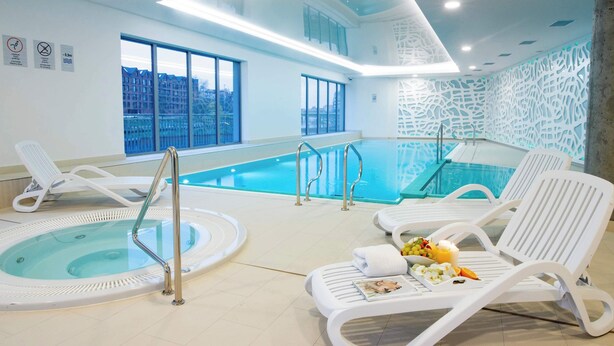 A modern indoor pool area with lounge chairs, a hot tub, and large windows offering city views.