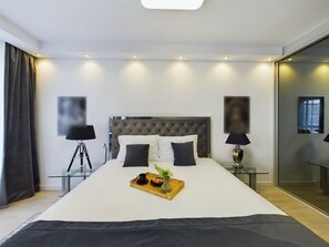 Un dormitorio de hotel moderno con una cama grande, lámparas de noche, una mesita de vidrio con bandeja y un espejo grande.
