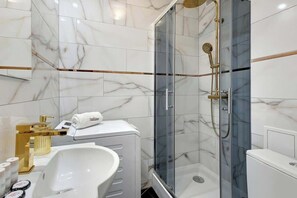 Ett modernt badrum med en dusch som är avskärmad med glasväggar, vita marmorplattor, en vit tvättställ och ett duschmunstycke i guld.