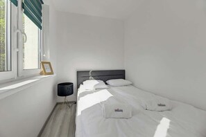 En säng för en person med vita sängkläder, en sänglampa och ett fönster med persienner.