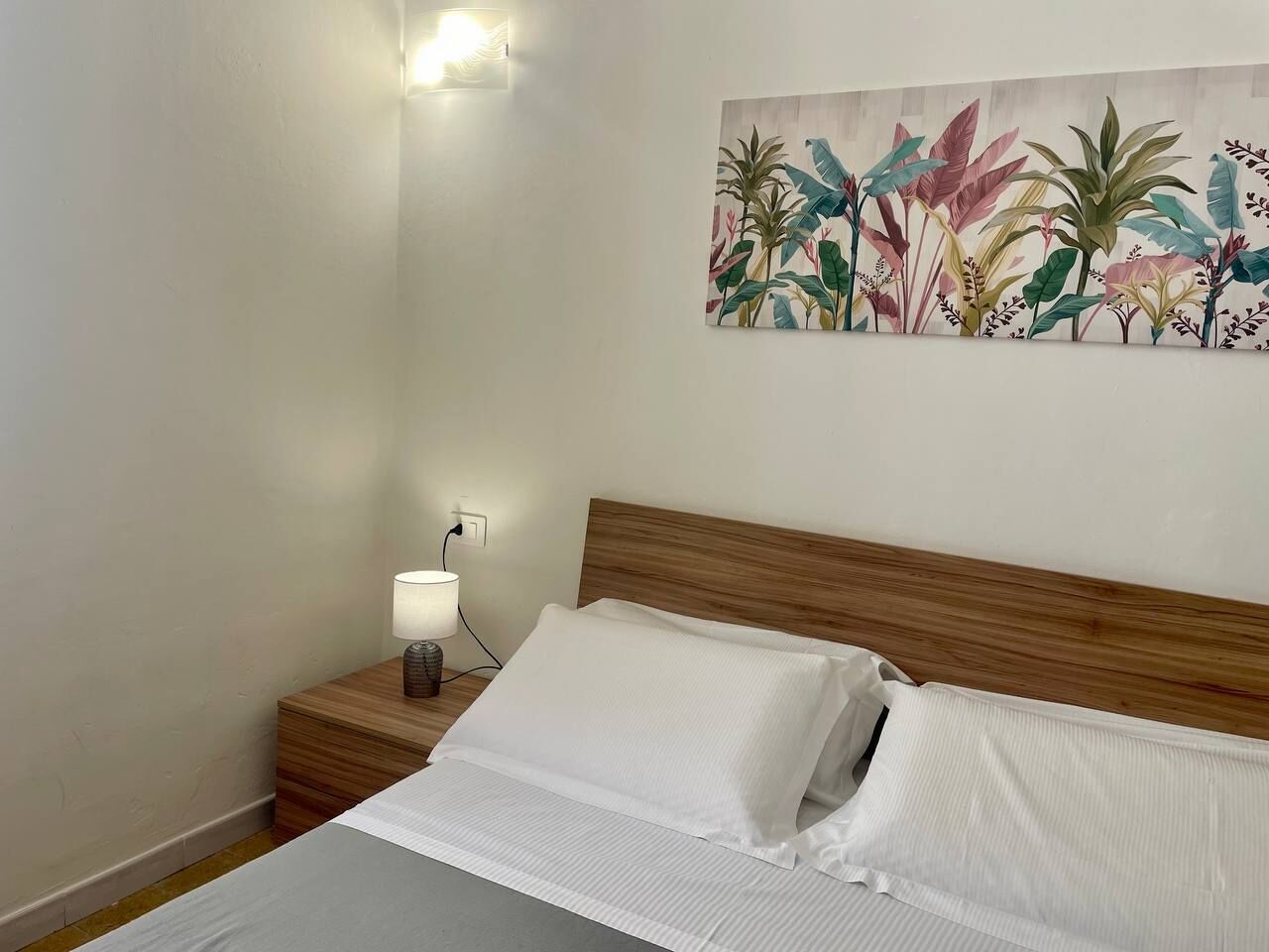 A neatly made bed with white linens, a wooden headboard, a bedside lamp, and a wall-mounted painting of tropical plants.