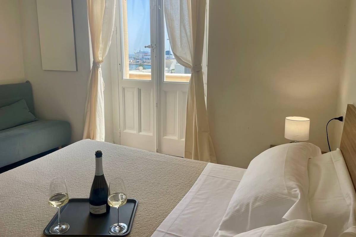 Quarto de hotel com cama, mesa de cabeceira com abajur, garrafa de vinho e duas taças em um tabuleiro.