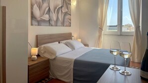 Una cama bien hecha con una colcha azul, una mesita de noche con una lámpara y dos copas de vino sobre una mesa.