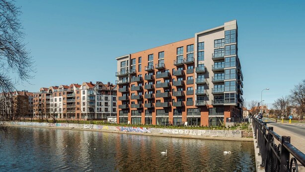 A modern apartment building with balconies, a river in the foreground, and a clear sky.