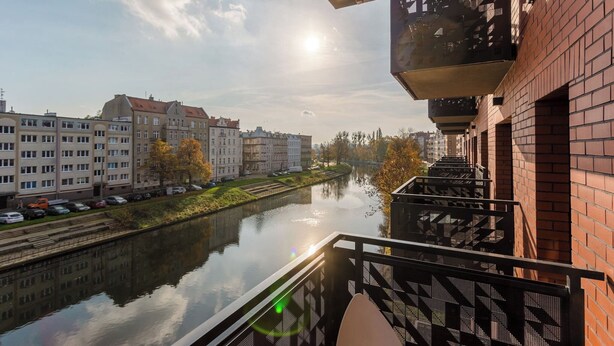 Une rivière, avec des bâtiments d’un côté et un balcon doté d’une rambarde en métal de l’autre.
