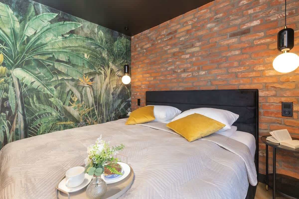 Ein Hotelzimmer mit Backsteinwand, einem großen Bett mit gelben Kissen, einem Wandgemälde von tropischer Vegetation und einem Nachttisch mit Lampe.