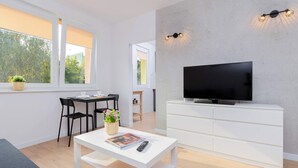 A modern living room with a flat-screen TV on a white cabinet, a dining table with chairs, and a small side table with a plant and a remote control. - Comfortable Studio | Close to the Beach and Reagan Park (Gdańsk)