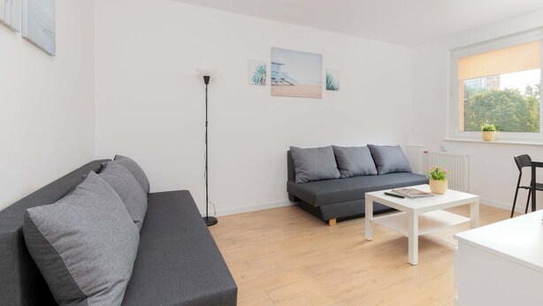 A modern living room with a grey sofa, a white coffee table, and a floor lamp. There are two paintings on the wall and a window with a view of trees. - Comfortable Studio | Close to the Beach and Reagan Park (Gdańsk)