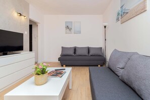 A modern living room with a flat-screen TV, grey sofas, a white coffee table with a magazine and flowers, and a small side table.