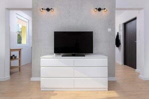 A modern living room with a flat-screen TV on a white cabinet, two wall-mounted lights, and a wooden table with a pot.