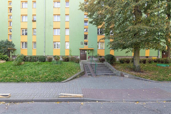 A multi-story building with a green and yellow facade, a central entrance with a staircase, and a tree in the foreground.