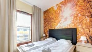 A hotel room with a bed, a nightstand, a window with curtains, and a wall with a tree and sun pattern. - Bright Apartment | River View and Parking Lot (Gdańsk)