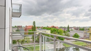 A balcony with a glass railing overlooking a cityscape with buildings and trees. - Bright Apartment | River View and Parking Lot (Gdańsk)