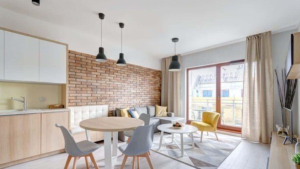 A modern kitchen with a brick accent wall, a dining area with a round table and chairs, and a living space with a sofa and yellow armchair. - Bright Apartment | River View and Parking Lot (Gdańsk)