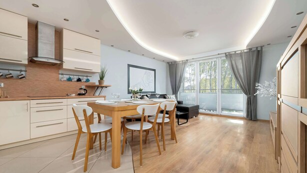 A modern kitchen with a dining area, wooden cabinets, and a large window with curtains.
