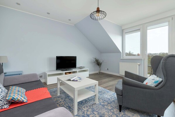 A modern living room with a flat-screen TV, a white coffee table, a grey armchair, and a sofa with patterned cushions.