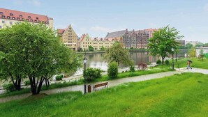 Un parc au bord de la rivière, avec un banc, des arbres et des bâtiments en arrière-plan.