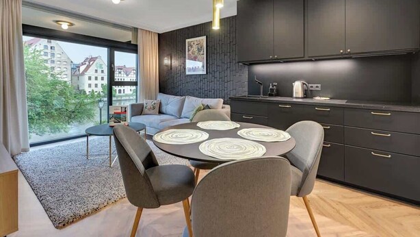 Una cocina moderna con armarios oscuros, un área de comedor con una mesa redonda y sillas, y un amplio ventanal con vista a edificios.