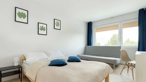 A bedroom with a bed, a bedside table, a chair, a window with curtains, and framed leaf artwork on the wall.