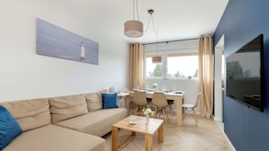 A modern living room with a sofa, a wooden coffee table, a dining area with chairs, a TV mounted on the wall, and a painting of a lighthouse.